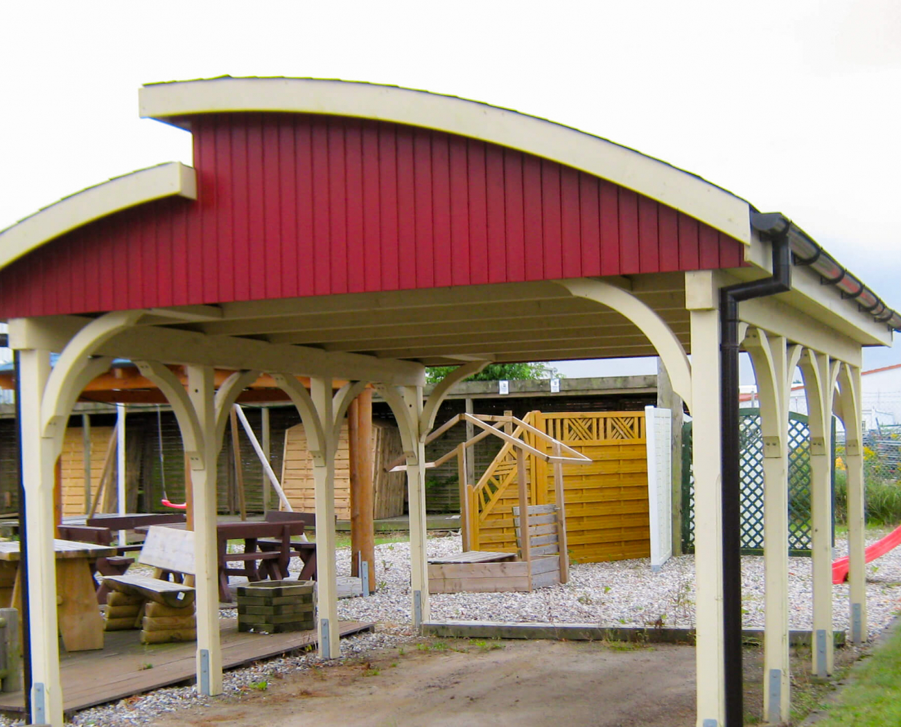 Symbolbild "Carport Holzschutz": Ein Carport mit einer außergewöhnlichen Dachform steht in einem Garten. Die Seitenteile des Dachs sind rot angestrichen. Der Carport selbst ist in einem hellen Holzton gehalten.
