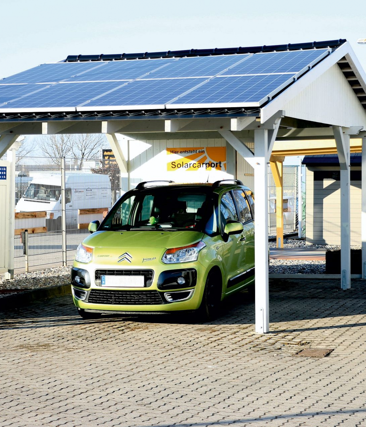 Ein weißer Solar Carport steht auf grauen Pflastersteinen. Auf dem Dach befinden sich acht Solarpanels, jeweils vier pro Reihe. Unter dem Solar Carport ist ein grünes Auto geparkt.