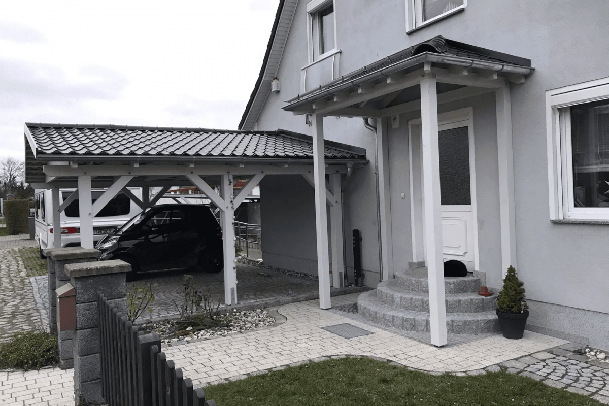 Seitenansicht eines Doppelcarports aus Holz, das mit dem grau-weißen Design zum angrenzenden Wohnhaus passt. Auf einem der beiden Parkplätze des Carports steht ein schwarzes Auto.