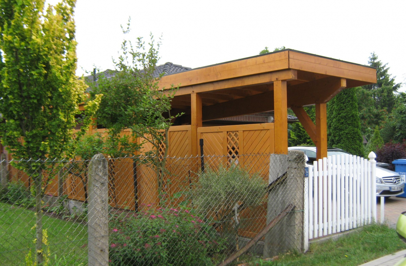 Ein schmaler Carport aus Holz grenzt an einen Gartenzaun. Der Carport besitzt ein flaches Dach. Im angrenzenden Garten sind einige Bäume, Pflanzen und Sträucher zu sehen.