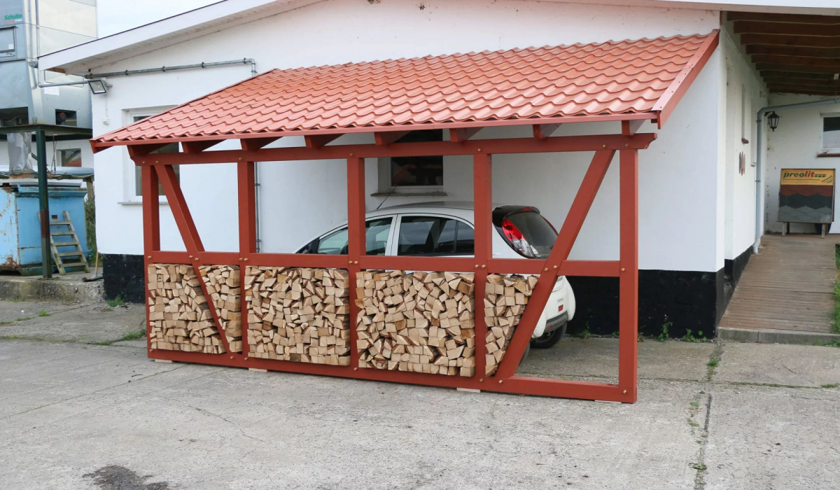 Ein Carport im Fachwerkstil wurde als Anbau direkt neben einem Haus platziert. Mehrere Holzscheite sind an den Wänden des Carports befestigt, während ein weißes Auto unter dem Carport parkt.