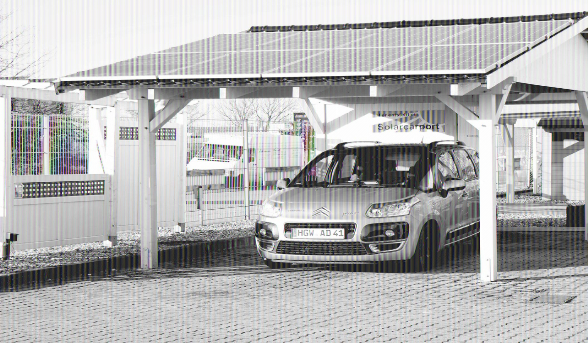 Ein grüner PKW parkt unter einem Carport mit Satteldach, auf dem mehrere Solarpanels installiert wurden. Der Carport ist aus weiß lackiertem Holz gefertigt und besitzt dunkle Dachziegel.