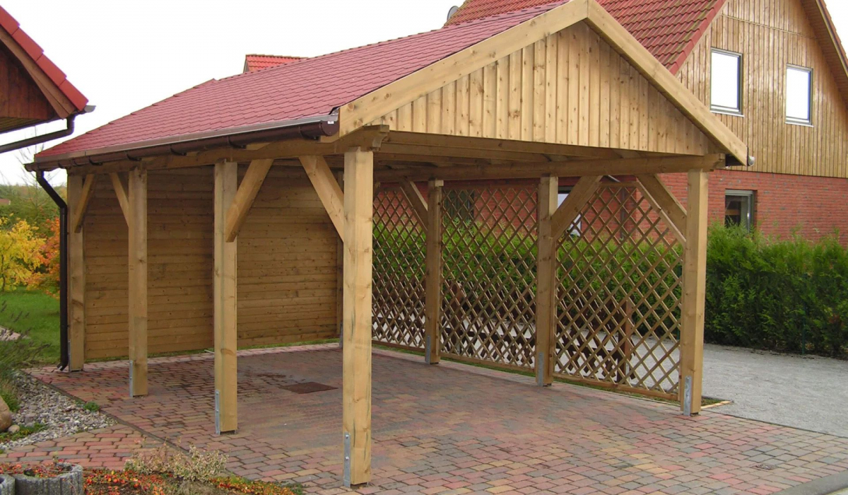 Elegantes Carport aus Holz mit einem Satteldach und roten Dachziegeln. Die Rückseite des Carports ist vollständig mit Holz verkleidet, während die angrenzende Wand mit einem Zaun versehen ist.