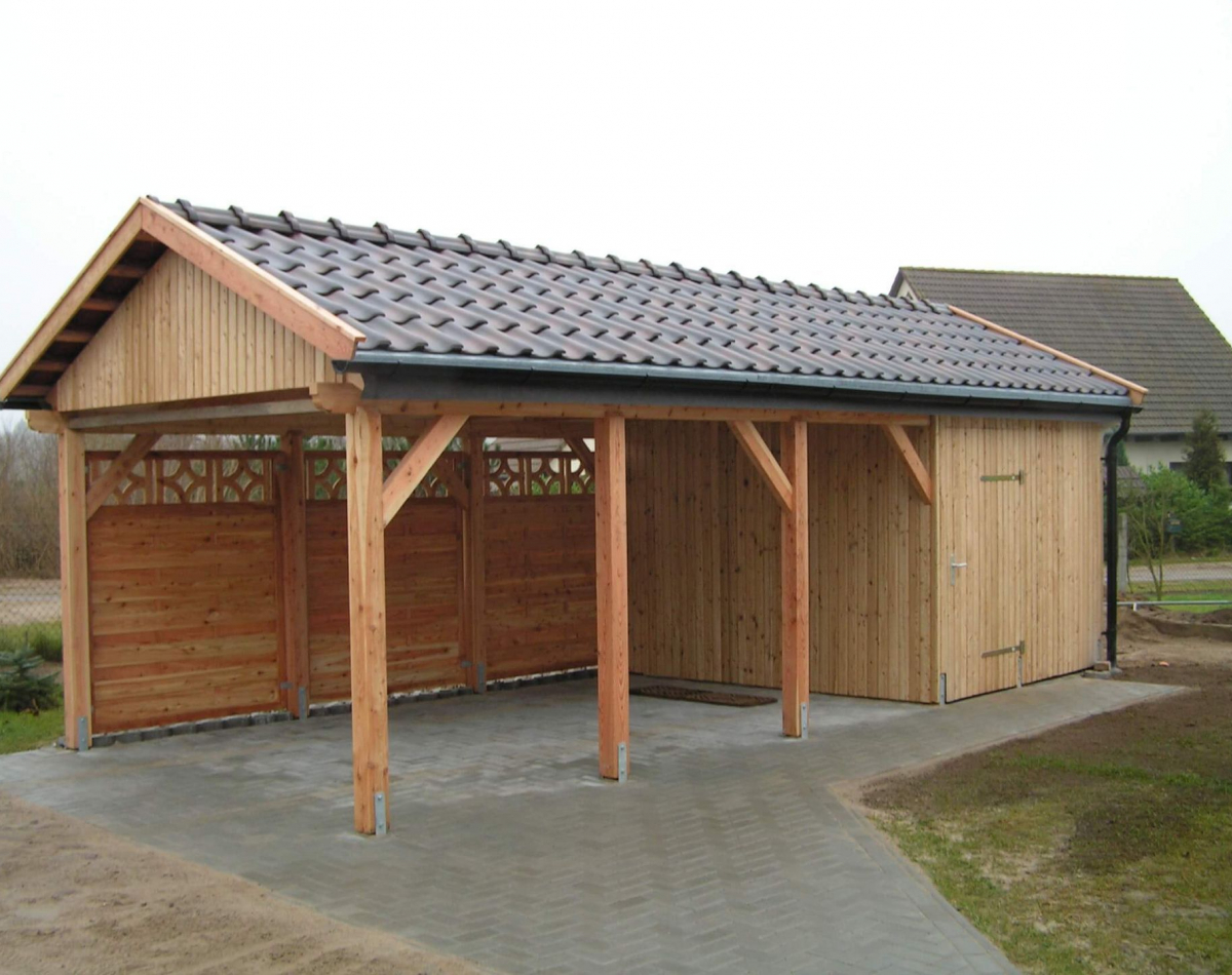 Ein Carport mit Satteldach steht auf gepflastertem Boden in einem Garten. Das typische Dach ist mit dunklen Dachziegeln ausgestattet. Der hintere Teil des Carports verfügt über einen kleinen Geräteschuppen.